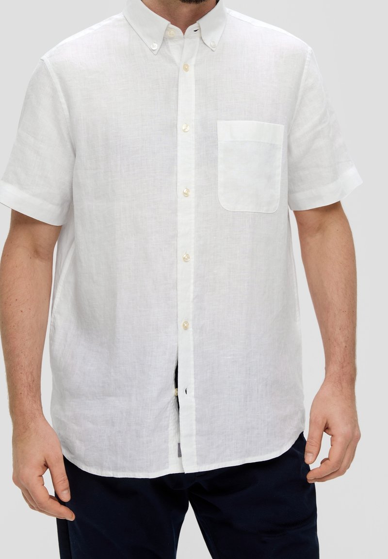 Man in een witte blouse met korte mouwen en een borstzakje, met marineblauwe broek, tegen een effen achtergrond.