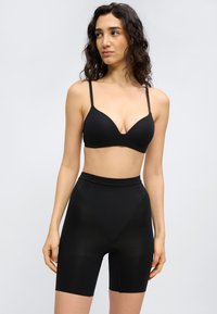 Γυναίκα με μακριά σγουρά μαλλιά που φοράει μαύρο σουτιέν και μαύρο σορτς με ψηλή μέση, στέκεται μπροστά σε απλό ανοιχτό φόντο.