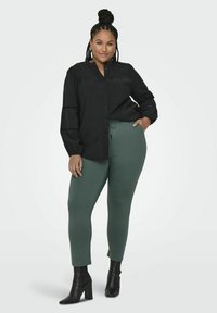 Svart bluse med knapper og puffede ermer, kombinert med grønne, tettsittende bukser og svarte høye støvler. Glatte teksturer, avslappet design.