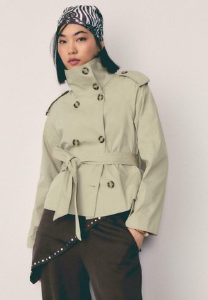 Młoda kobieta w jasnobeżowym trenczu z paskiem, ciemnych spodniach i czarno-białej chustce na głowie, stojąca na jednolitym tle.