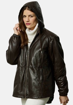 Frau trägt eine dunkelbraune Lederkapuzenjacke über einem weißen Rollkragenpullover und hält die Kapuze mit ihrer linken Hand.