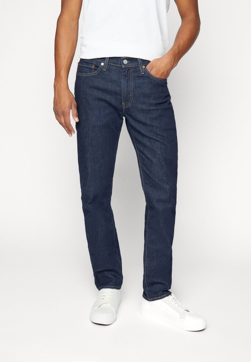 Vaqueros de mezclilla azul oscuro con un corte recto, con costuras naranjas y cinco bolsillos, combinados con una camiseta blanca y zapatillas blancas.