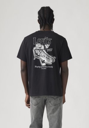 Osoba u crnoj Levi's majici s crtanim hot dogom koja sjedi na stolici, a na stražnjoj strani piše "Keeping your buns toasty".