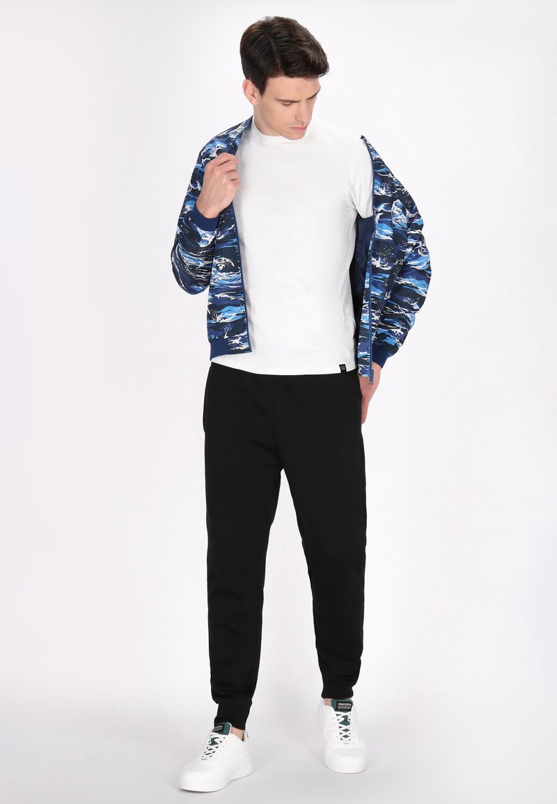 Blauwe en witte geblokte bomberjack over een wit T-shirt, gecombineerd met zwarte joggingbroeken en witte sneakers. Eenvoudige, casual sportkleding.