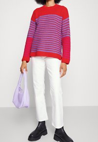 Maglione a righe rosse e viola con maniche lunghe, abbinato a pantaloni bianchi a zampa d'elefante e stivali neri chunky. Tiene una borsa lilla.