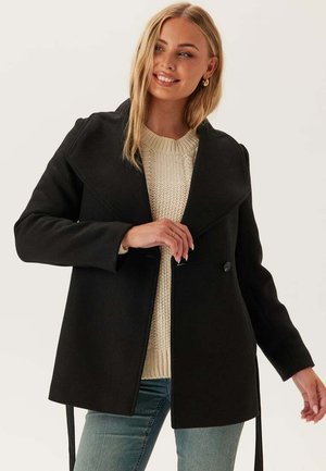 Manteau en mélange de laine noir avec un large col, fermeture à double boutonnage et liens de ceinture. Associé à un pull en tricot crème et à un jean bleu.