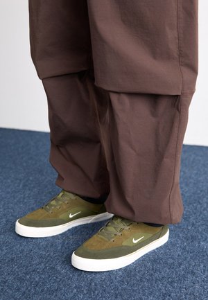 Zaļi un pelēki Nike sporta apavi ar baltām detaļām, ar teksturētu augšdaļu un šņorēšanu. Valkāti ar brūnām vaļīgām biksēm uz zilā paklāja.