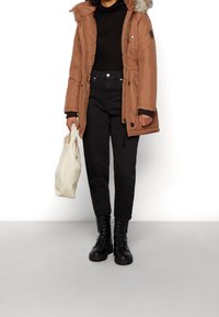 Brun isoleret parka med pelsforet hætte, sort højhalset trøje, højtaljede sorte bukser og sorte snørebåndsstøvler; holder en cremefarvet mulepose.