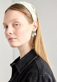 Schwarze Jeansjacke mit einem weichen grünen Stoffhaarband. Silberne skulpturale Ohrringe mit fließendem Design, Seitenansicht einer Person.