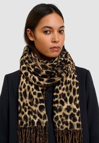 Sciarpa con stampa leopardata in tonalità nera e marrone, texture morbida, rifinita con frange lunghe, indossata sopra un giubbotto scuro, sfondo neutro.