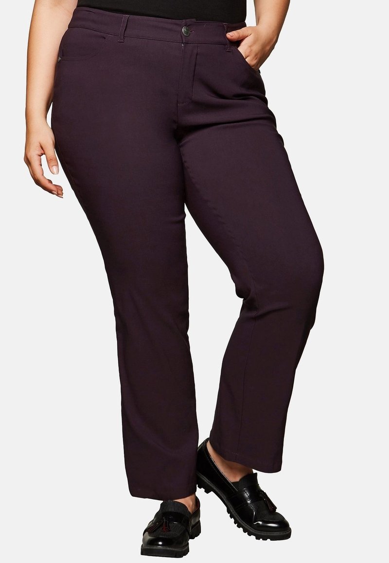 Personne aux formes courbes portant un pantalon droit violet foncé, des mocassins noirs et un haut noir, avec une main dans la poche et l'autre détendue.