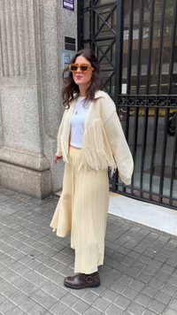 Krēmo adīta kardigāna ar fringām, balts tops, dzeltenas plakanas platībās bikses un brūni potītē ievelkami zābaki; papildināts ar dzeltenām saulesbrillēm.