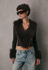 Maglione marrone corto con texture soffice attorno al collo e ai polsini, abbinato a jeans di denim blu chiaro a vita alta. Occhiali da sole scuri.