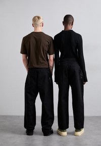 Dos modelos de espaldas, uno con una camiseta marrón y pantalones negros, y el otro con una blusa de canalé negra y pantalones negros, ambos usando zapatillas.