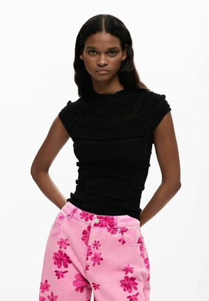 Frau trägt ein eng anliegendes schwarzes Kurzarmshirt kombiniert mit hoch taillierten pinkfarbenen Hosen, die mit großen dunkelrosa Blumenmustern verziert sind.
