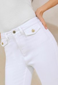 Vaqueros de mezclilla blanca de cintura alta con herrajes dorados, que presentan dos bolsillos frontales y una textura suave. Primer plano del detalle de la cintura.