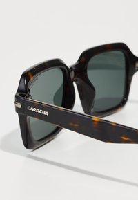 Ochelari de soare tortoise cu lentile pătrate, cu nuanță verde, având un logo metalic Carrera pe brad și un finisaj lucios.