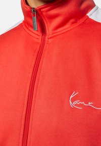 Chaqueta roja con cremallera y collar alto, con acentos en blanco en los hombros y un logotipo bordado en blanco en el lado izquierdo. Textura de tela suave.