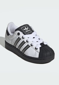 Čierno-biele tenisky s textúrovaným koženým zvrškom, tromi čiernymi pruhmi a gumovou špičkou. Majú biele šnúrky a branding na jazyku.