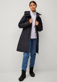 Zwarte lange jas met een capuchon, voorzien van ritssluiting en drukknoopsluitingen, zijzakken, gedragen over een lichtgrijze fleece top en blauwe jeans, gecombineerd met zwarte laarzen.