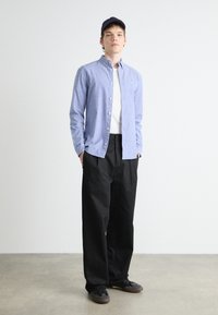Joven de pie con una gorra negra, camisa de botones azul claro sobre una camiseta blanca, pantalones anchos negros y zapatos negros sobre un suelo gris.