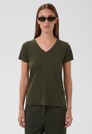 Femme aux cheveux bruns raides portant des lunettes de soleil noires étroites, un t-shirt vert foncé à col en V et manches courtes, et un pantalon sombre, debout devant un fond uni.
