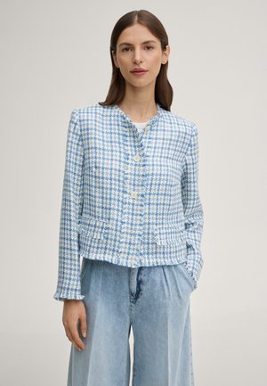 Vrouw draagt een blauw-wit geruit jasje met knopen en franjeranden, gecombineerd met lichtblauwe, wijde, hooggesneden jeans.