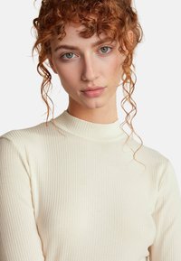 Gestreiftes, creamfarbenes Langarmshirt mit hohem Kragen. Es hat eine glatte Textur und eine enganliegende Silhouette, die eine dezente Kurvenform an den Ärmeln zeigt.
