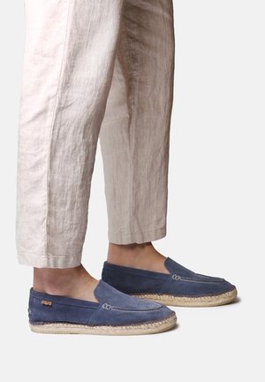 Noge v svetlo bež lanu hlačah in modrih semišastih espadrilkah z pletenim jute podplatom na svetli podlagi.