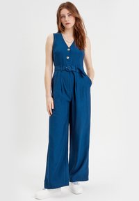 Un vestito blu senza maniche con una vita aderente, gambe larghe, dettagli con bottoni e una cintura coordinata. Texture del tessuto liscia. Indossate sneakers bianche.