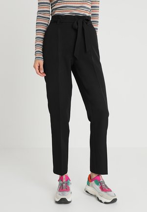 Mujer con pantalones negros de cintura alta con lazo en la cintura, camiseta de manga larga a rayas y zapatillas de deporte gruesas y coloridas sobre un fondo liso.