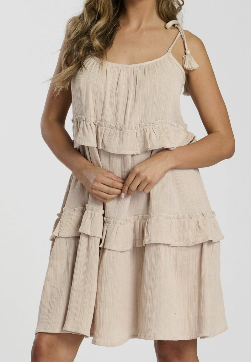 Femme portant une robe beige sans manches avec des volants et des bretelles à nouer, tenant le tissu au niveau de la taille avec les deux mains.