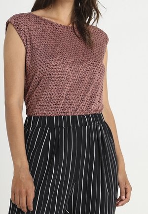 Vrouw draagt een mouwloze bruine top met kleine zwarte driehoeken en een zwarte broek met hoge taille met witte verticale strepen.