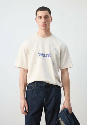 Hombre con una camiseta blanca con el texto "Want", vaqueros azul oscuro, sosteniendo una chaqueta vaquera doblada en su mano izquierda contra un fondo liso.