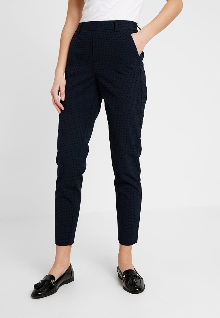 Femme portant un pantalon bleu marine slim à petits pois noirs, longueur cheville, un haut blanc sans manches et des mocassins noirs, les mains dans les poches.
