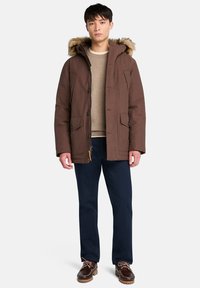 Brun parkajacka med foder av päls, framfickor och knäppning. Bärs över en beige tröja, ihop med marinblå byxor och skor.