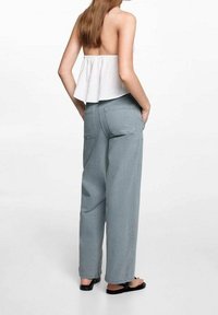 Vrouw met lang haar draagt een witte strapless top, lichtblauwe wijde broek en zwarte sandalen, staat met haar handen in de zakken, met haar rug naar ons toe gekeerd.