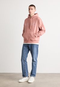 Hoodie in hellrosa mit weißem "HUGO"-Logo, mit einer frontalen Kängurutasche, darüber ein weißes T-Shirt getragen, kombiniert mit blauen Jeans und weißen Sneakern.