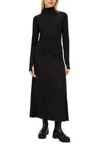 Robe maxi noire à manches longues avec un col montant, en tissu texturé avec de subtils paillettes, portée avec des bottines noires.