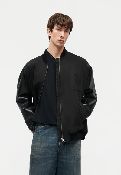 Jeune homme portant une veste bomber noire à manches en cuir, un t-shirt noir et un jean bleu ample, debout les mains dans les poches.