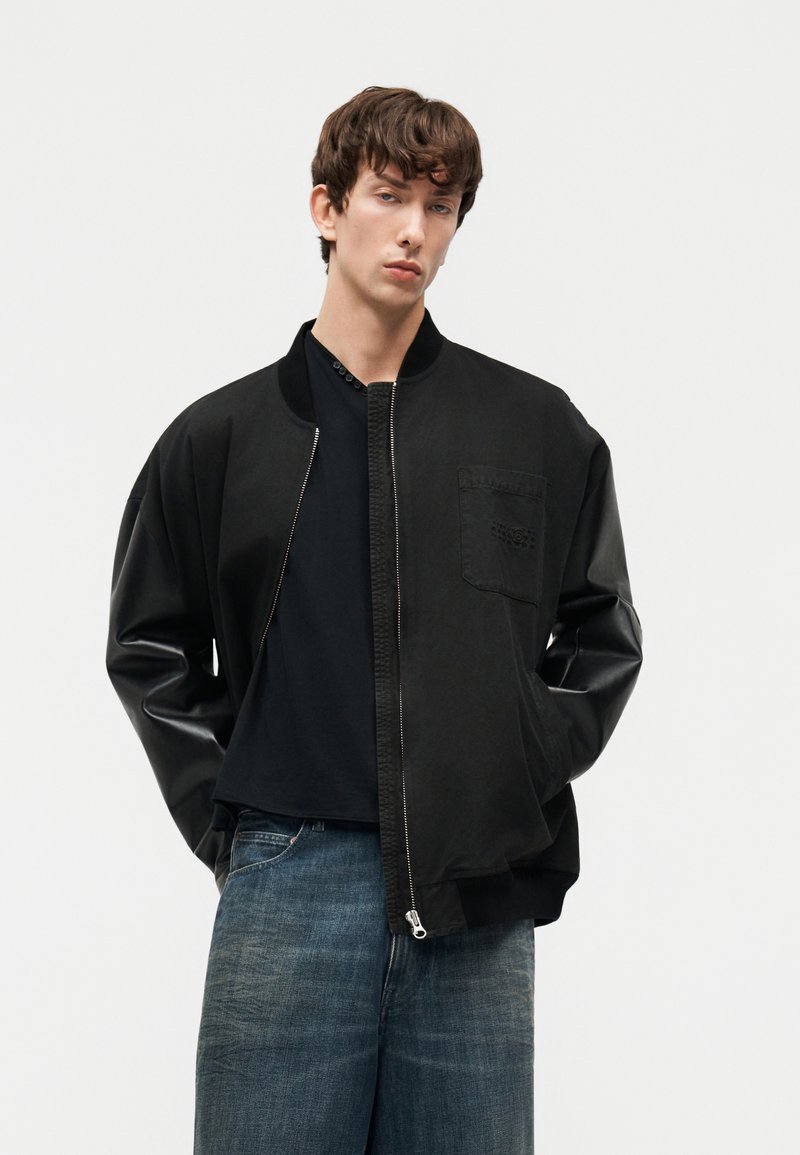 Jonge man die een zwart bomberjack met leren mouwen, een zwart T-shirt en losse blauwe jeans draagt, staand met handen in de zakken.