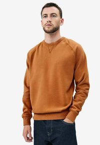 Sudadera de algodón marrón con cuello redondo y puños de canalé, que presenta una superficie texturizada y un corte relajado. Se lleva con unos vaqueros de mezclilla oscuros.