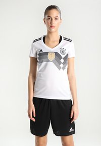 Witte sporttrui met zwarte strepen, met een centraal embleem en logo, gecombineerd met zwarte shorts. Gemaakt van ademend materiaal.