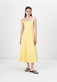 Vestido midi de lino amarillo con escote cuadrado y tirantes gruesos. Falda en forma acampanada y diseño minimalista, combinado con chanclas negras.