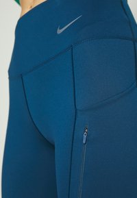 Zblízka modré vysoké sportovní legíny s boční kapsou na zip a reflexním logem Nike na boku.