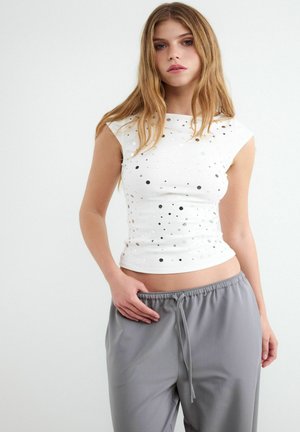 Junge Frau mit langen Haaren, die ein weißes ärmelloses Top mit runden Ausschnitten und graue Hose mit Kordelzug trägt, steht mit einer Hand in der Tasche.