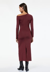 Frau, die mit dem Rücken zur Kamera steht, trägt ein langes burgunderfarbenes Kleid mit einem schulterfreien Ärmel und schwarze, hochhackige Stiefeletten auf weißem Hintergrund.