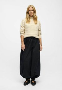 Frau mit langen blonden Haaren, die einen beigefarbenen Strickpullover, lockere schwarze Jogginghose und schwarze Schuhe trägt, steht vor einem schlichten weißen Hintergrund.