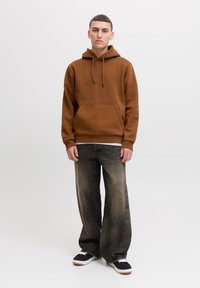 Ung mand står mod en hvid baggrund iført en brun hættetrøje, slidte vide jeans og sorte sneakers med hvide såler.