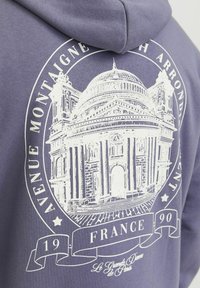 Lila kapucnis felső, amelyen egy történelmi épület fehér grafikája látható, körbevéve kör alakú szöveggel, amely tartalmazza a "Avenue Montaigne", "Franciaország" és "1990" feliratokat.